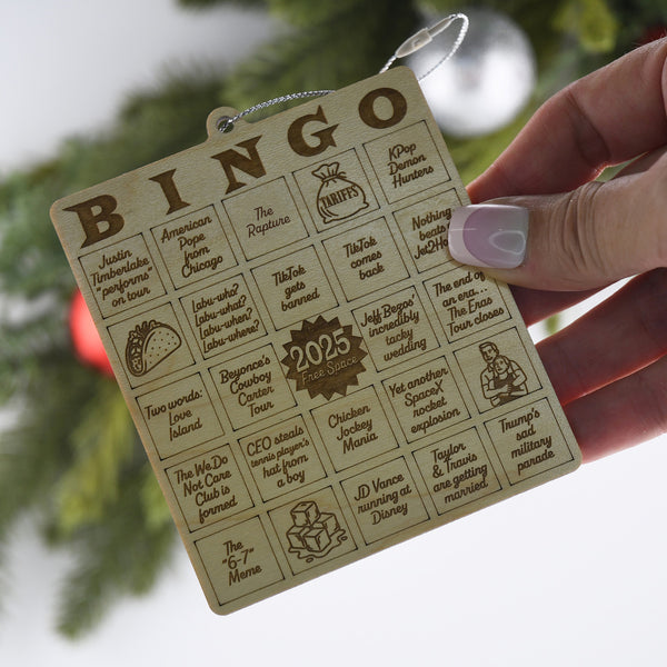 Wooden bingo card with humorous text and 2023 design, held by a hand against a blurred green background.