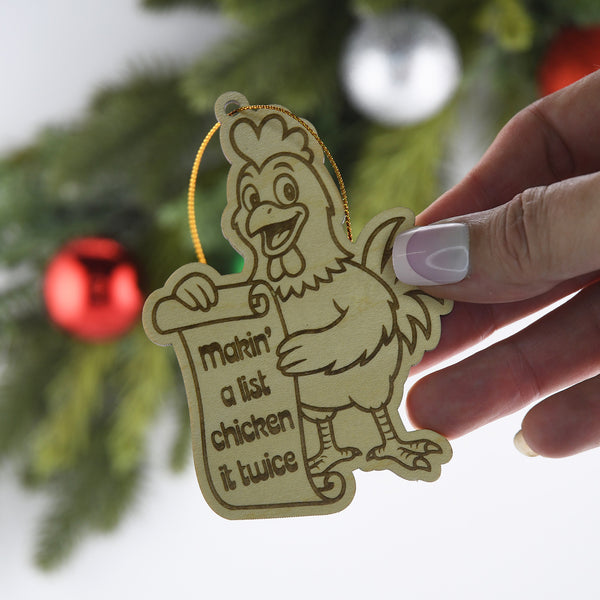 Decorative ornament with a chicken holding a sign, held by a hand against a Christmas tree background.