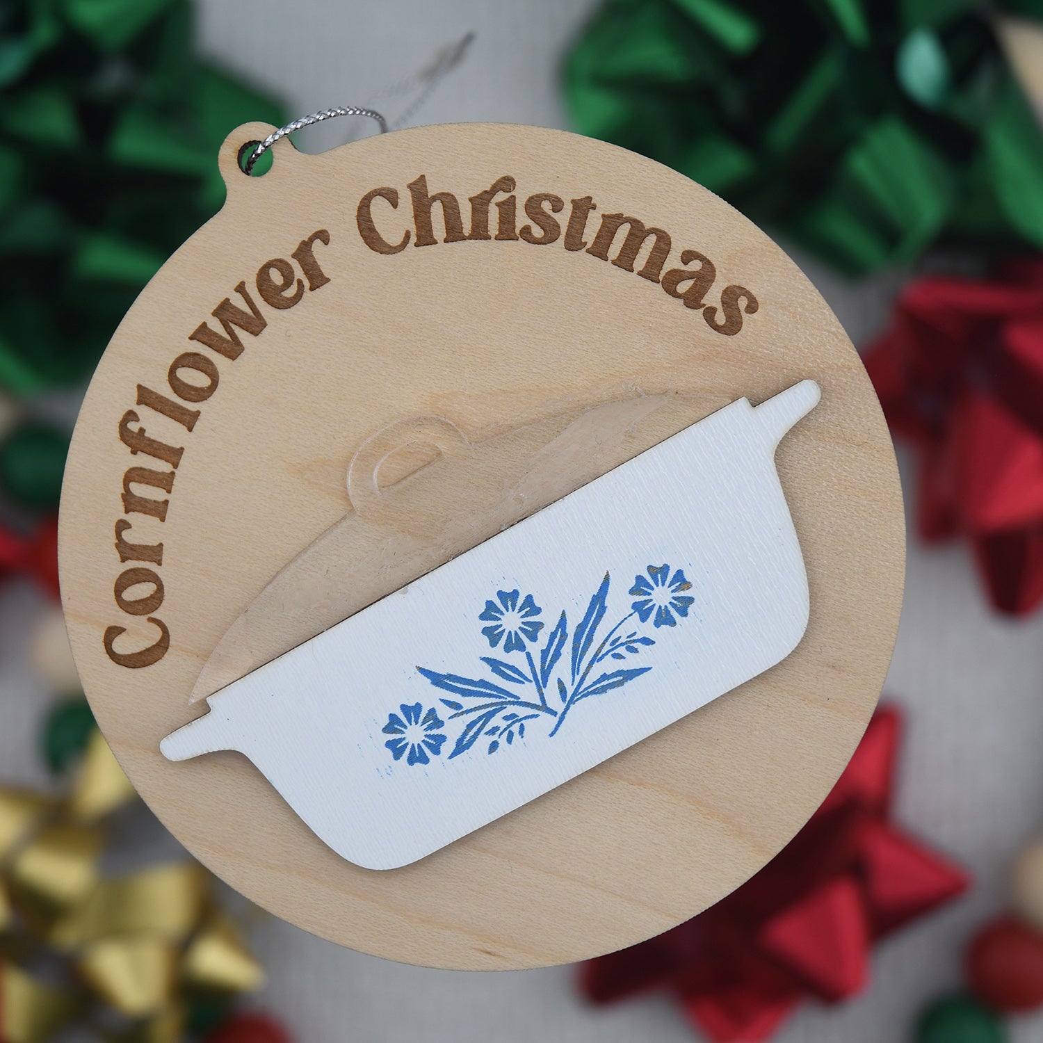Wooden Christmas ornament with 'Cornflower Christmas' text and floral design on a blurred festive background.