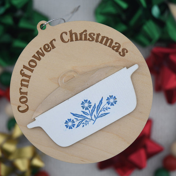 Wooden Christmas ornament with 'Cornflower Christmas' text and floral design on a blurred festive background.