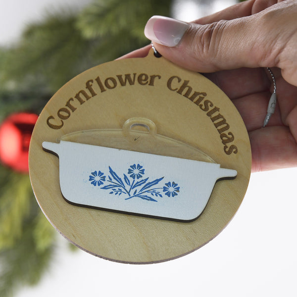 Decorative ornament with 'Cornflower Christmas' text and floral design, held by a hand against a blurred green background.