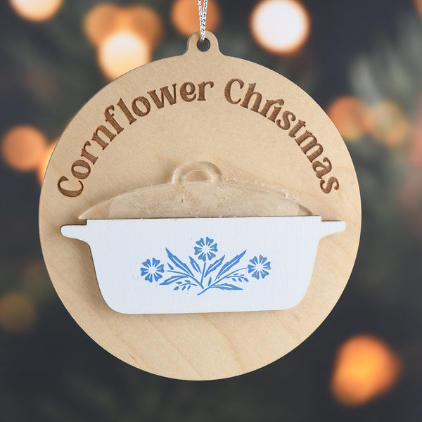 Wooden Christmas ornament with 'Cornflower Christmas' text and floral design on a blurred lights background
