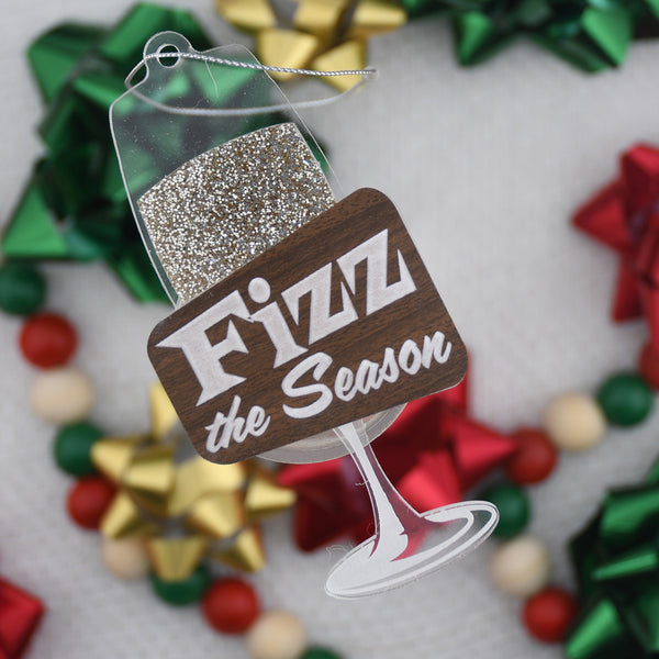 Decorative ornament shaped like a wine glass with 'Fizz the Season' text against a blurred Christmas bow background