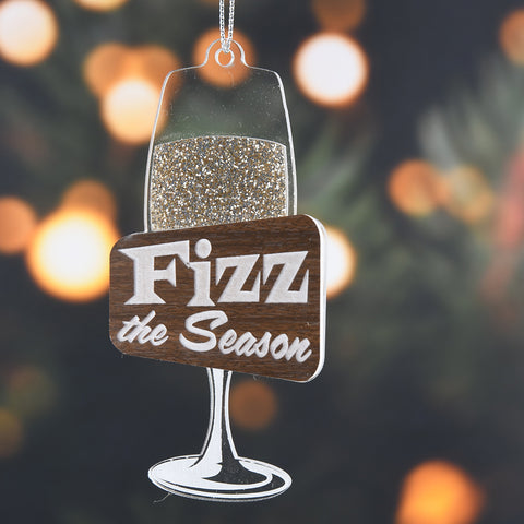 Decorative ornament shaped like a wine glass with 'Fizz the Season' text against a blurred Christmas tree background.