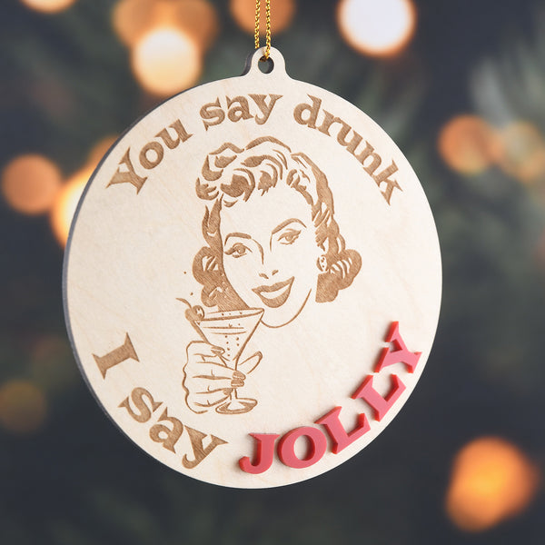 Wooden ornament with a woman holding a martini and text 'You say drunk, I say Jolly' hanging in front of a tree background