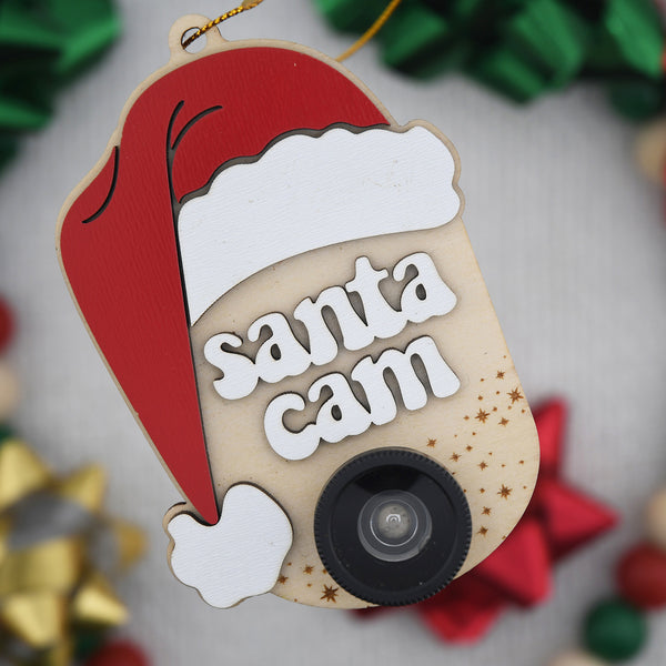 Ornament with a Santa hat, the words "Santa Cam" and a plastic camera lens hanging in front of a blurred background