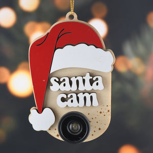 Ornament with a Santa hat, the words "Santa Cam" and a plastic camera lens hanging in front of a blurred background