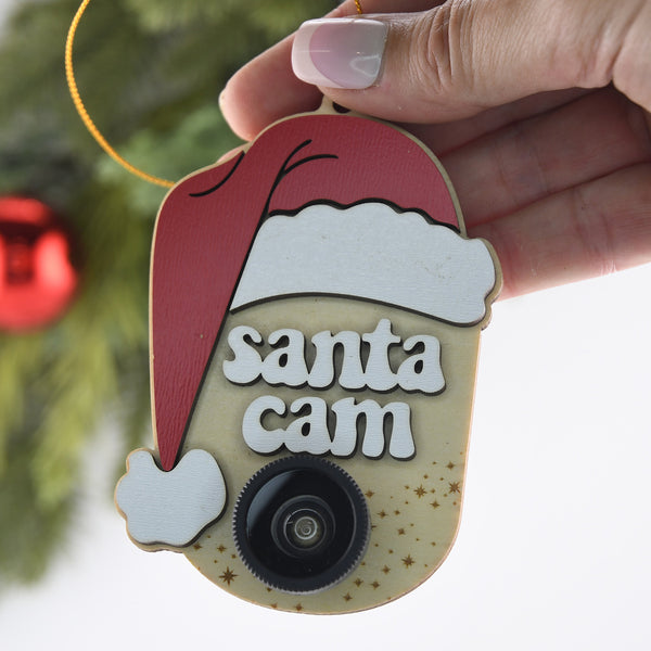 Ornament with a Santa hat, the words "Santa Cam" and a plastic camera lens being held by a woman's hand.