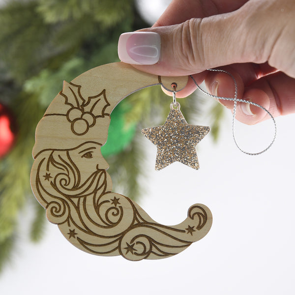 Wooden moon ornament with star decoration held by a hand against a blurred Christmas tree background.