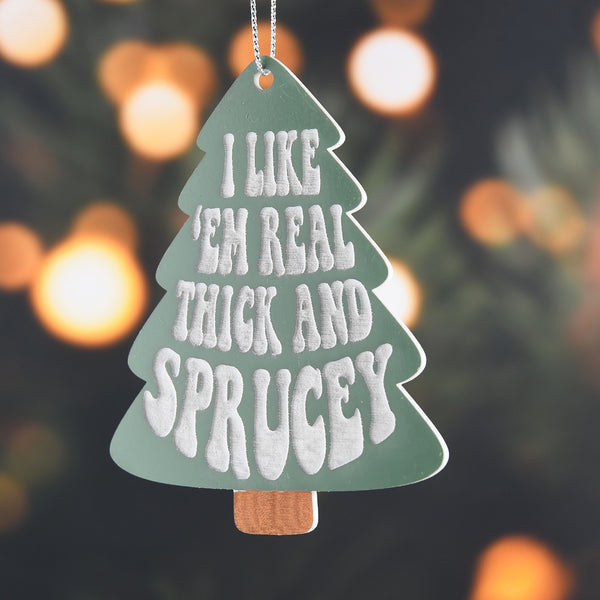 I Like em Real Thick and Sprucey ornament hanging in front of blurred tree background
