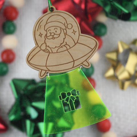 Decorative ornament with Santa Claus in an UFO and a green base with a gift icon, against a blurred Christmas bow background.